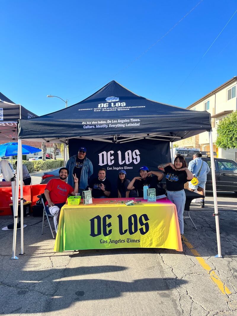 Photo of people standing under a tent and by a table with a yellow tablecloth that reads 'De Los'