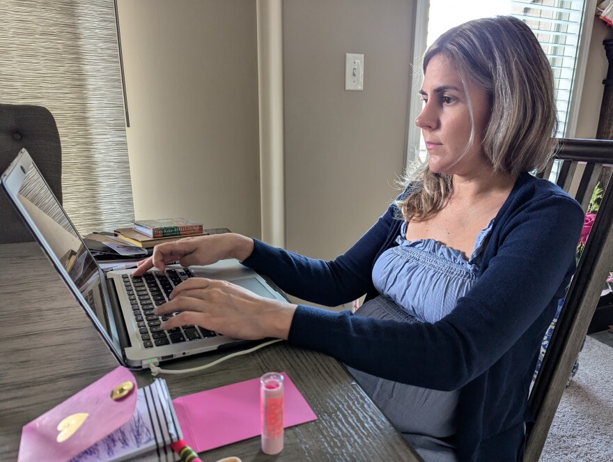 A woman sits at a dinner table inside her home. She is typing at a laptop computer.