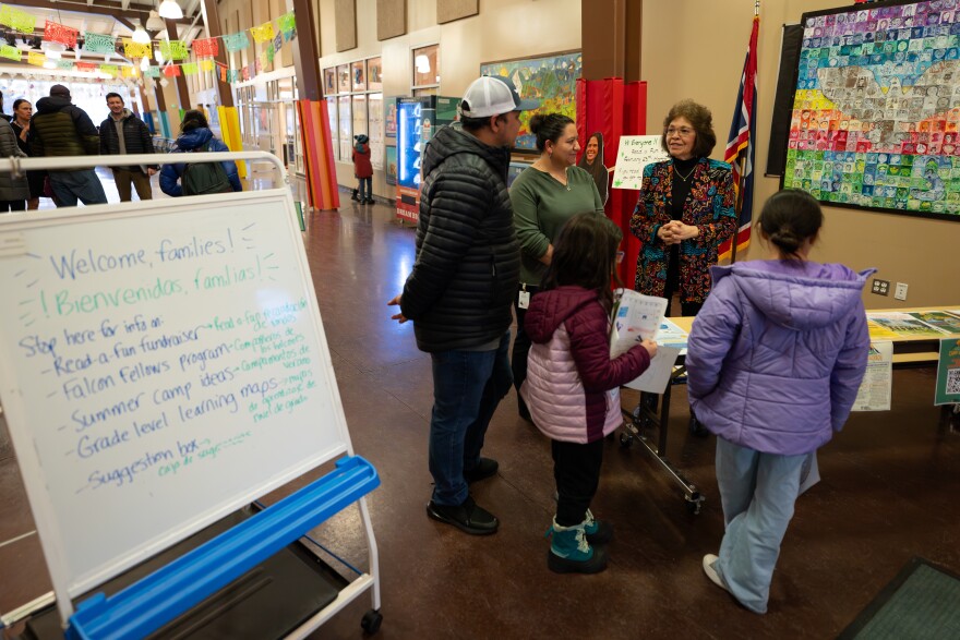 Bilingual signs in English and Spanish welcome families into a bright, colorful room. A Latino woman stands behind a table speaking to a family.