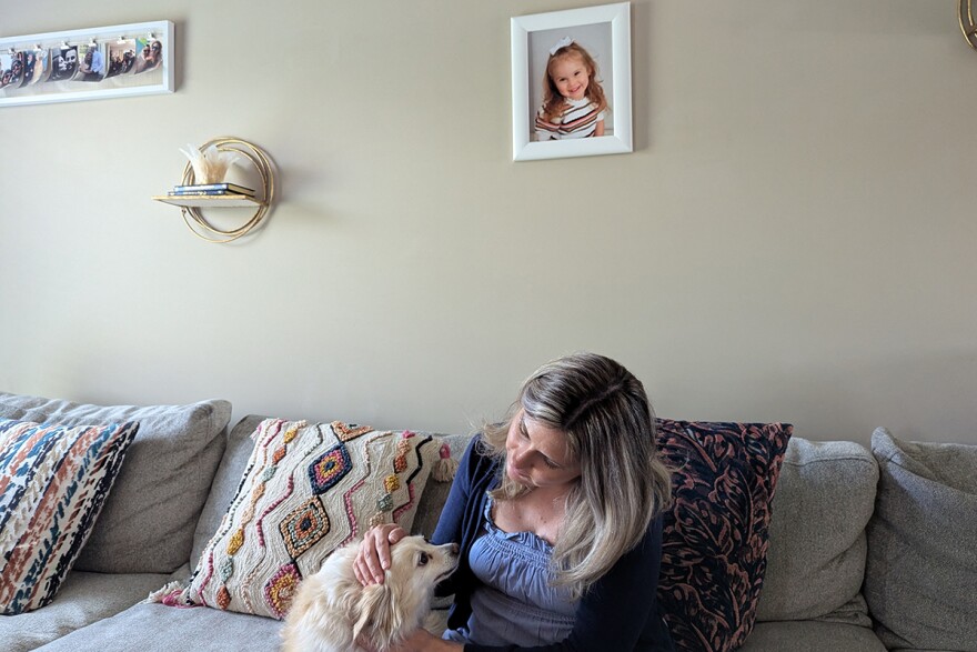 A woman stands inside her home near some shelves that display South American-themed dolls, family photos, and Christian collectibles.