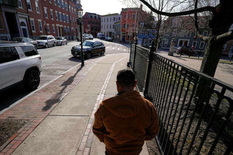 A man walks away from the camera along a sidewalk beside a fence, on a street lined with houses and parked cars.