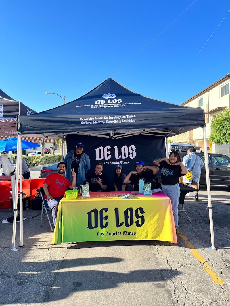 Photo of people standing under a tent and by a table with a yellow tablecloth that reads 'De Los'