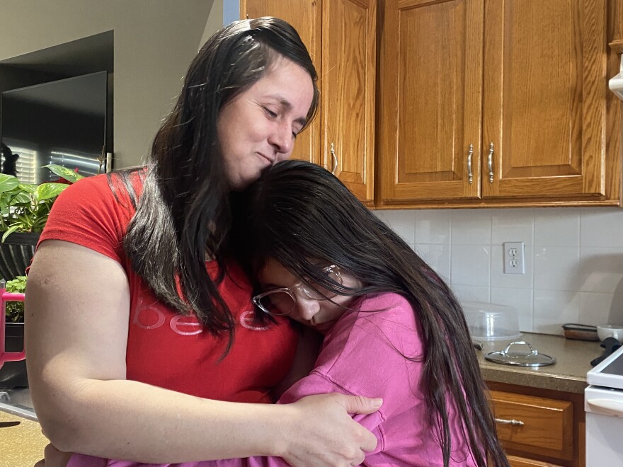 Woman in red T shirt and long black hair hugs little girl also with long black hair and a pink T shirt in kitchen