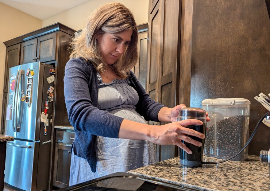 A woman stands at a granite kitchen counter. A refrigerator can be seen behind her. She is holding onto a coffee bean grinder and watching it grind coffee.