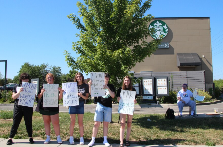 Five people stand in front of Starbucks holding signs against the company's alleged union busting. Another sits behind them.