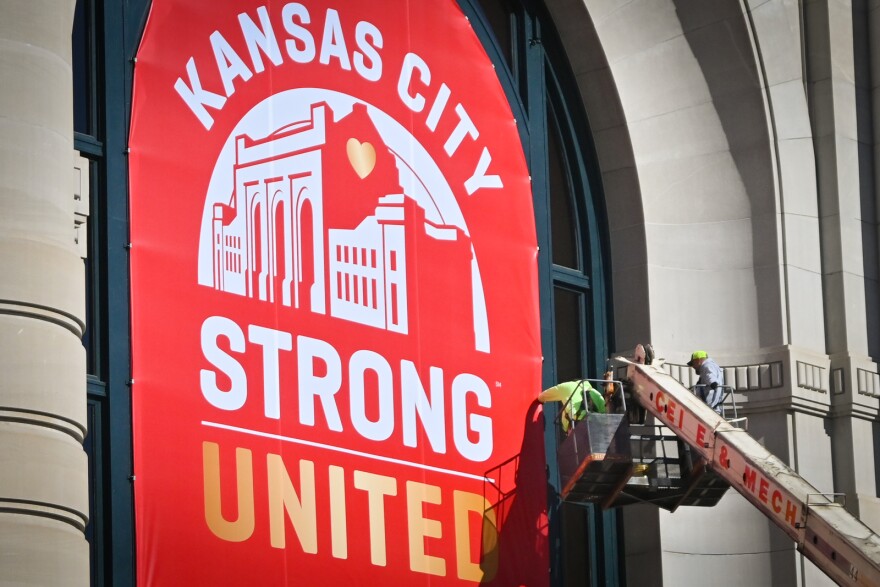 Men standing in a bucket that is raised by a crane work to hang a large red and white banner outside a stone building. The banner reads "Kansas City Strong United."