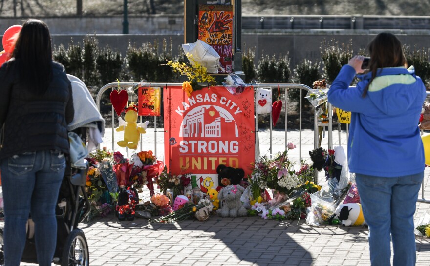 Outdoor photo in bright sunlight of a metal barrier that is decorated with red and yellow balloons. There are flowers gathered on the pavement in front of it along with small stuff animals. A sign attached to the metal barrier reads "Kansas city Strong, United." Two people can be seen with their backs to the camera. One is taking a photograph of the scene and the other is holding on to a baby stroller and looking at the display.