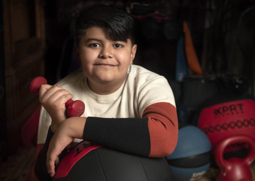 Child lifting a small red dumbbell in a home gym setting.