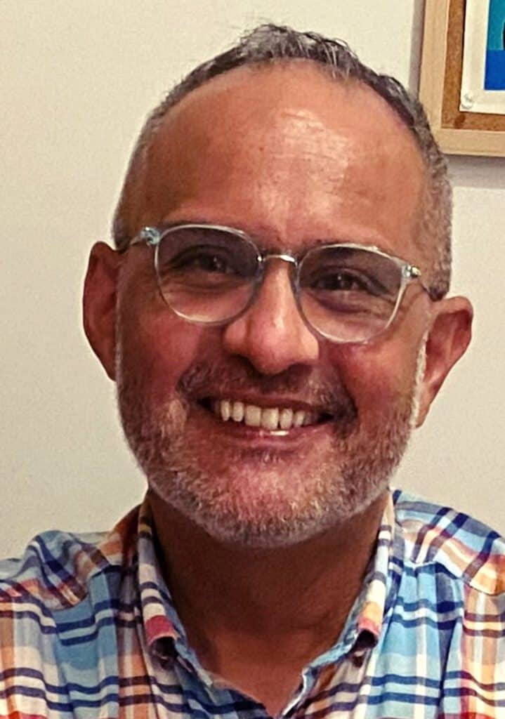 Latino man smiling, wearing glasses and a plaid shirt, in an indoor setting.