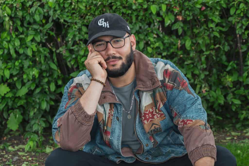 Latino man with glasses and beard sitting outdoors, wearing a cap and colorful jacket.