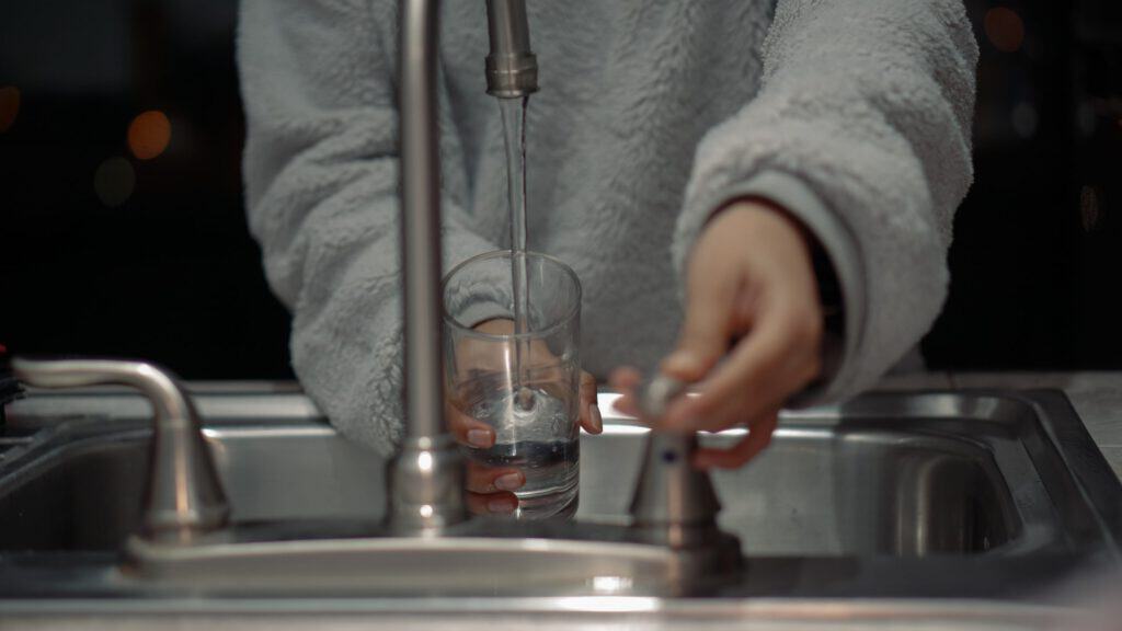 A person filling a glass with water from a kitchen sink faucet.