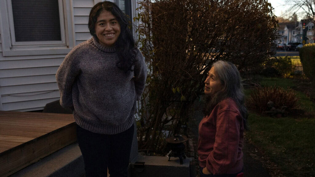 A woman and an elderly woman smiling outdoors in a suburban yard during fall.