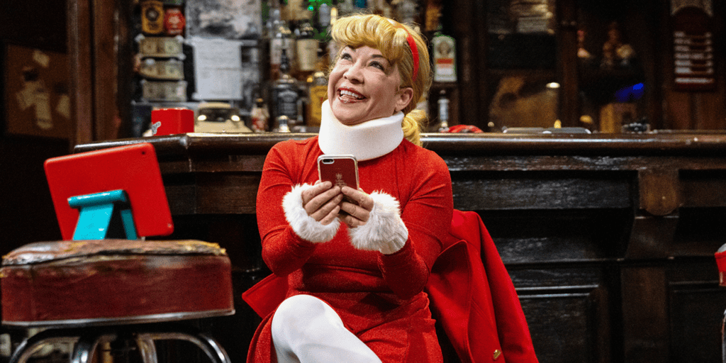 A festive woman dressed as Santa Claus smiling with a phone in a bar setting.