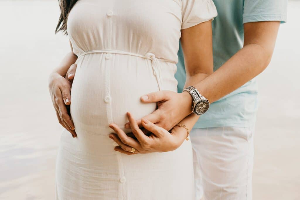 Pregnant woman with partner holding her belly at the beach, emphasizing support for Spanish-speaking.