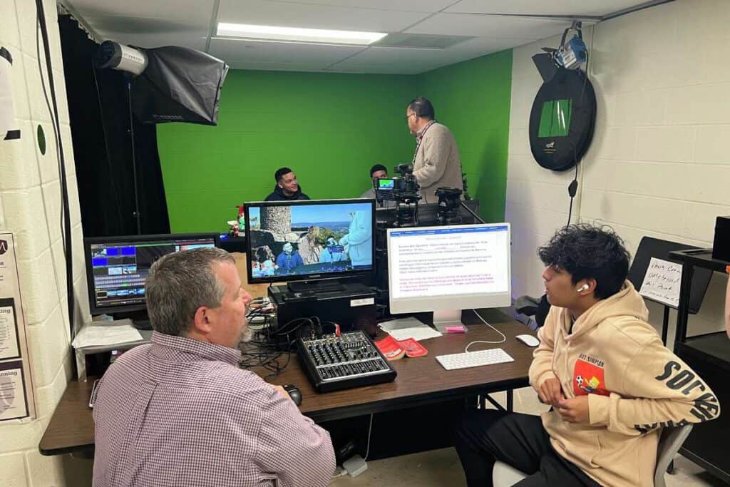 Students filming Spanish news broadcast in classroom with green screen.
