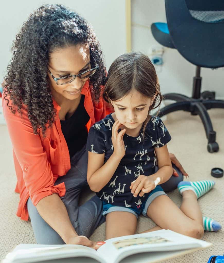 Latina foster mother reading a book to young girl in a supportive home setting.