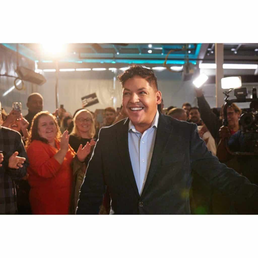 Latino candidate smiling at campaign event with supporters.