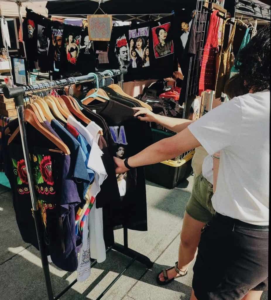 Shoppers browsing vibrant Latino clothing at Mercado market stall.