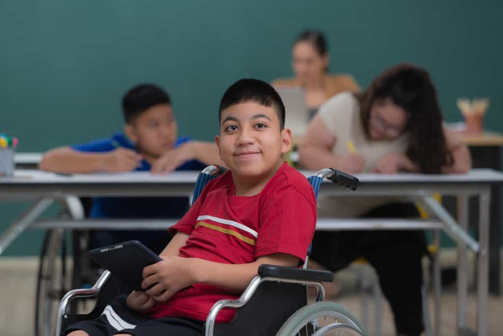 Accessible student in wheelchair smiling in classroom with teachers and classmates in background.