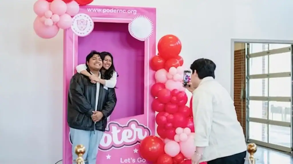 Two Latino women hugging at a voting-themed event with colorful balloons and a photo booth.