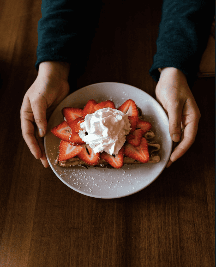 Strawberry waffle with whipped cream on a white plate, held by hands.
