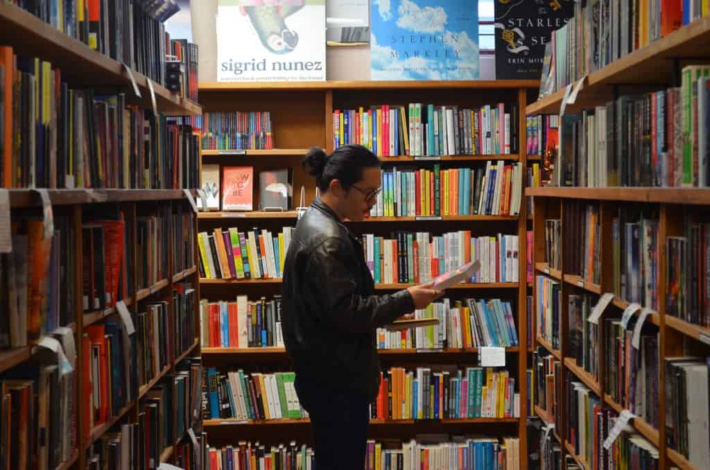 Woman reading a book in a bookstore aisle surrounded by colorful shelves.