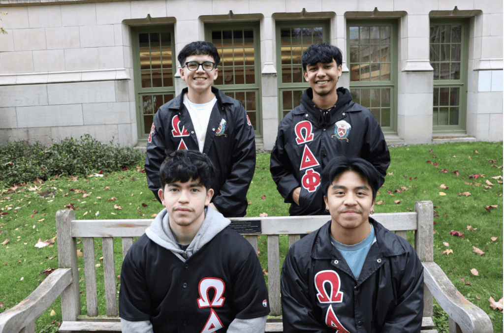 Four diverse students wearing fraternity jackets outdoors at university campus.