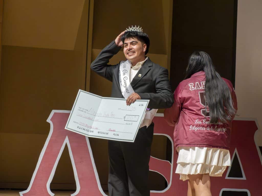 Young man wearing crown and sash holding a large check at a Latino celebration event.