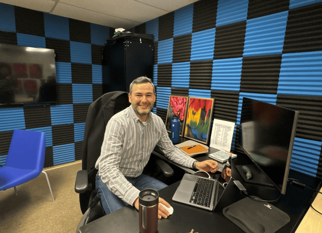 A man working at a desk with dual monitors in a soundproof recording studio.