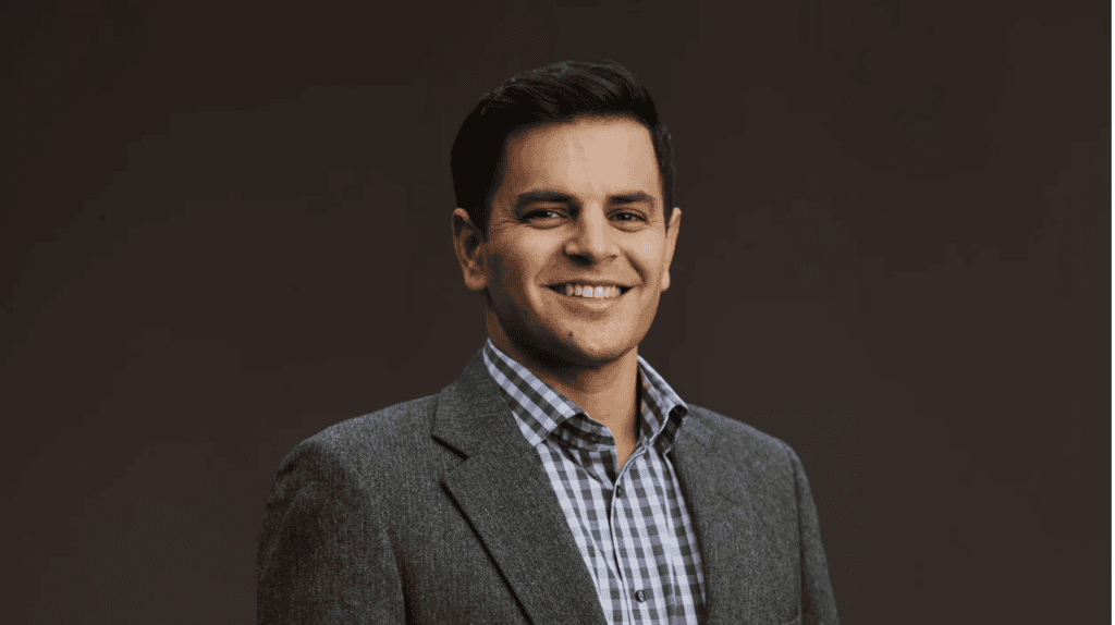 Professional Latino news anchor smiling in a gray suit and checkered shirt, representing Latino News.