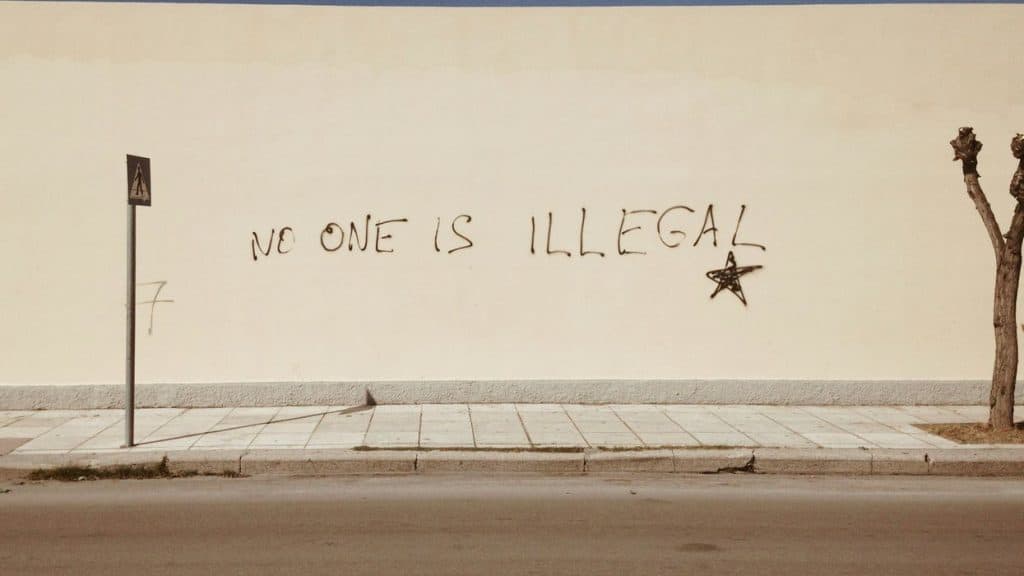 Graffiti message "No One Is Illegal" on a white wall in an urban setting.