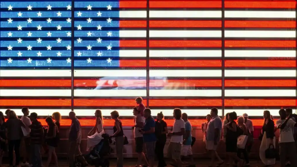 American flag illuminated with people walking in front.
