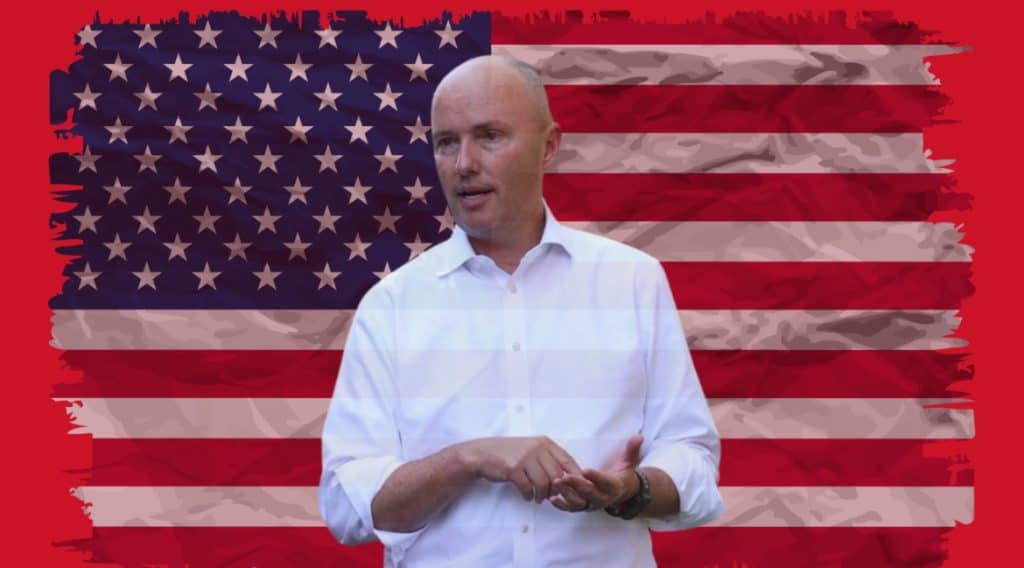 A man in a white shirt speaking in front of an American flag background.