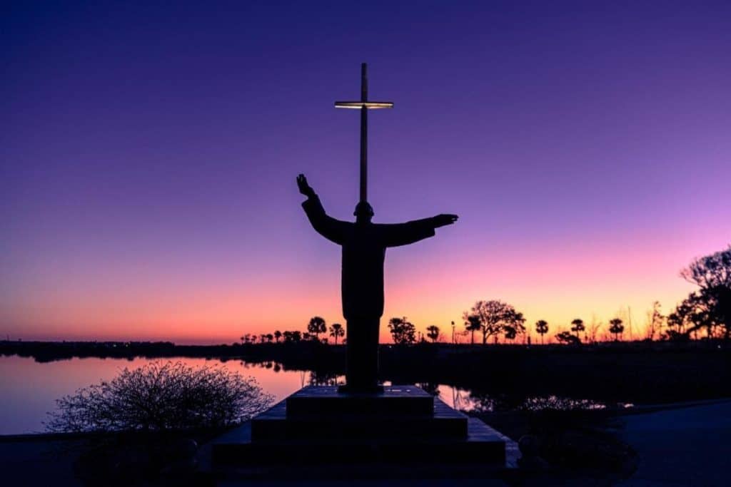 A person standing with arms outstretched in front of the Christ of the Redeemer statue at sunset in.