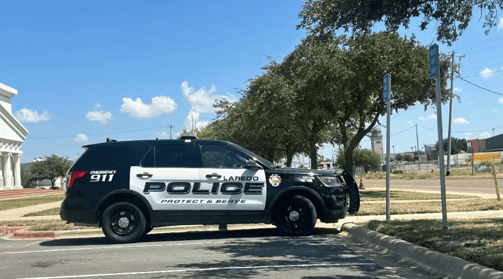 Laredo police vehicle parked near border crossing under blue sky and trees.