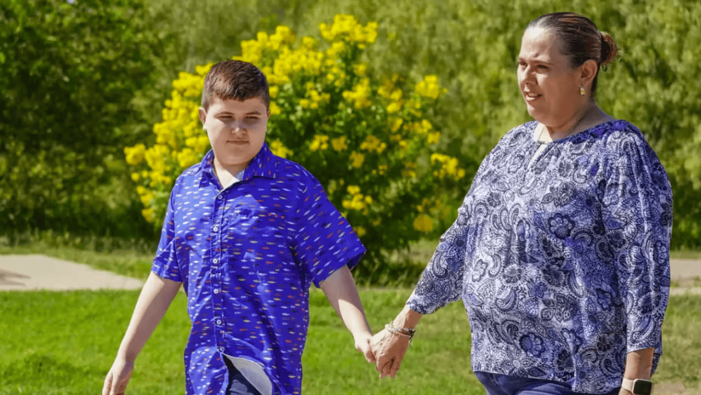 A boy and woman holding hands outdoors in a park with green trees and yellow flowers.