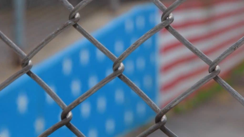 Venezuelan migrants behind a chain-link fence with a blurred flag in the background.