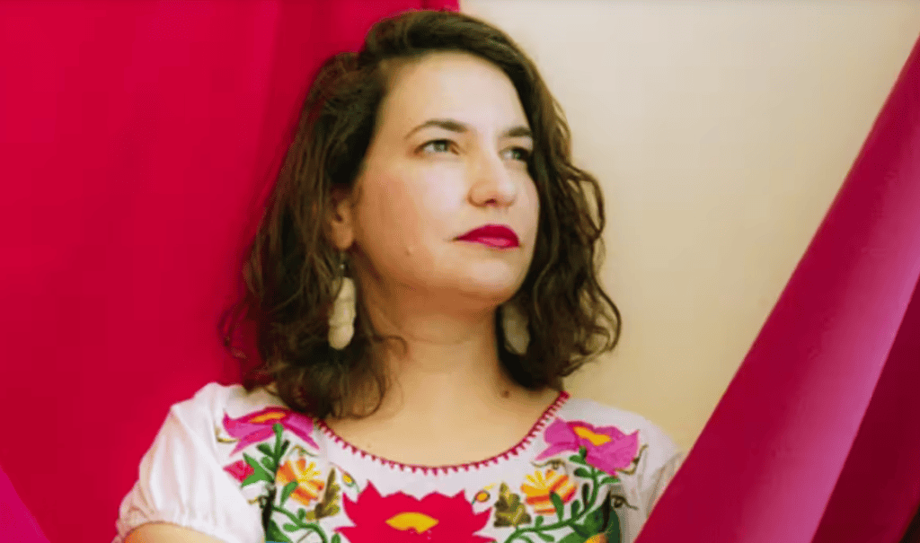 A woman with dark wavy hair and colorful embroidered blouse, holding a pink musical instrument, agai.