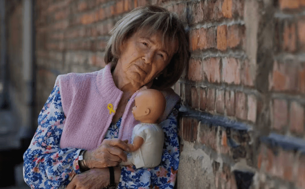A woman holding a doll near a brick wall, reflecting resilience and remembrance.