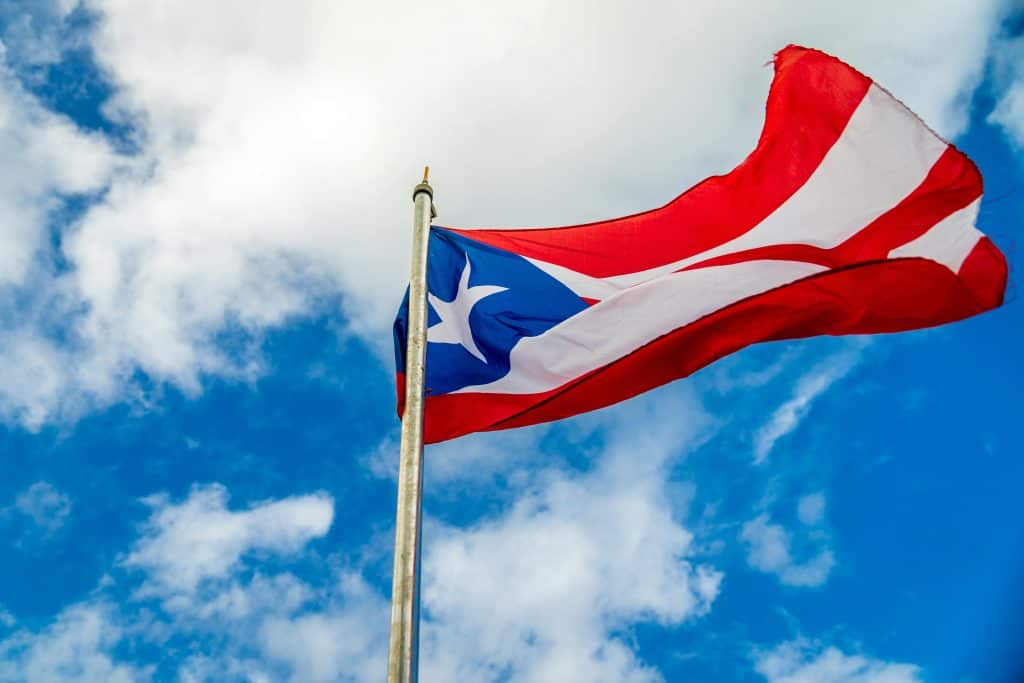 A vibrant Puerto Rican flag waving on a flagpole against a bright blue sky with clouds.