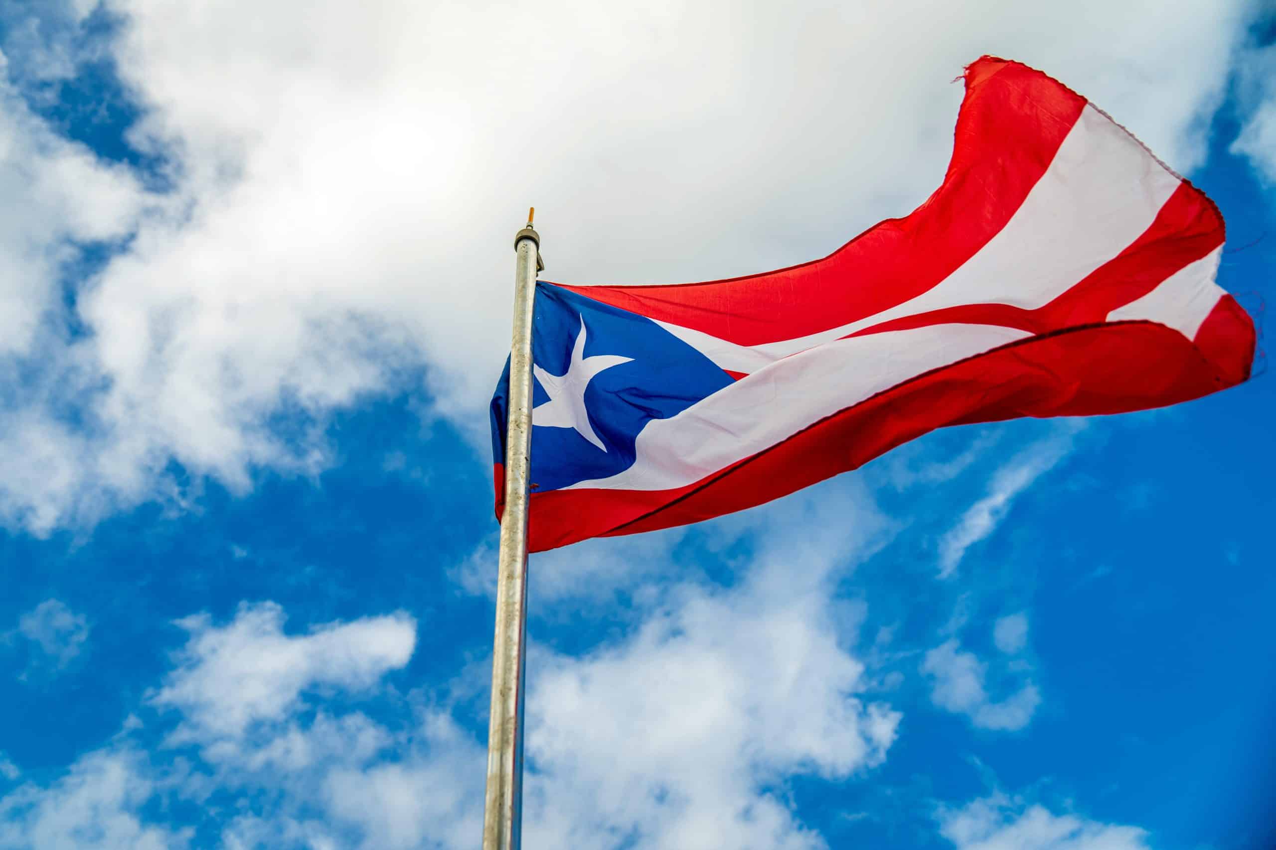 A vibrant Puerto Rican flag waving on a flagpole against a bright blue sky with clouds.