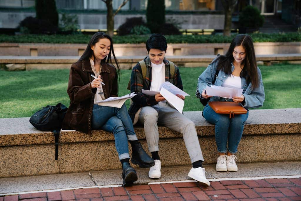 Three students studying together on a bench in a university campus setting outdoors.