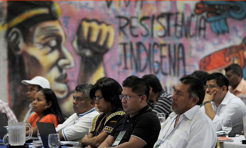 Diverse group of people attending a Latino news conference with activist mural in background.