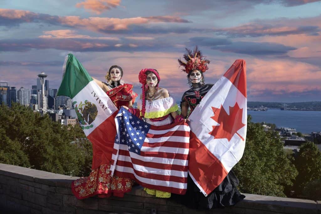 Three women in traditional Mexican and Canadian costumes holding flags with city skyline background.