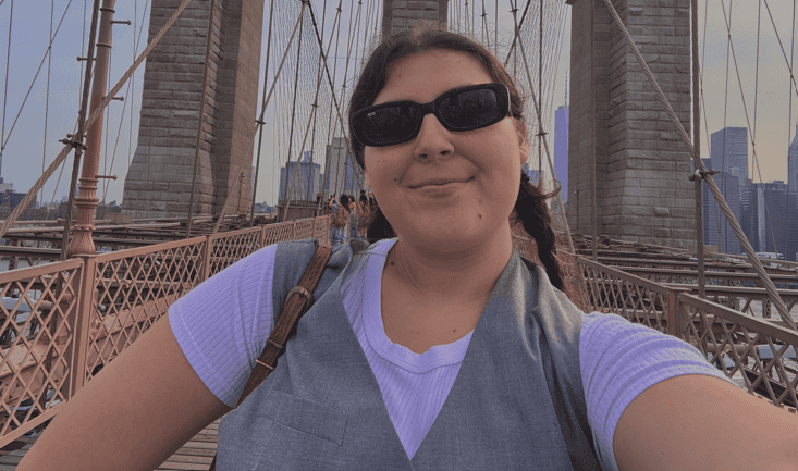Woman taking a selfie on Brooklyn Bridge with city skyline in background.