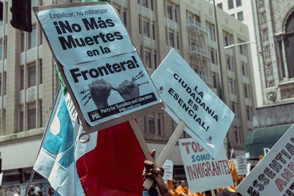Protesters hold signs against border deaths and militarization in an urban setting.