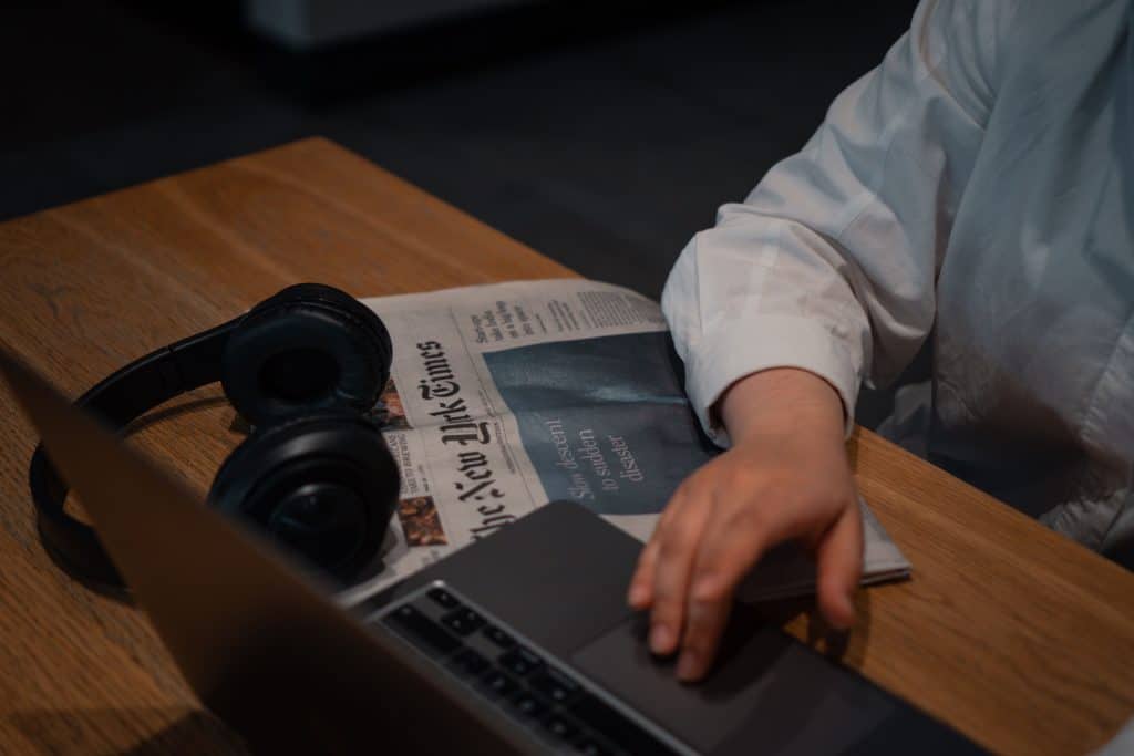 Person working on laptop with newspaper and headphones on wooden table.