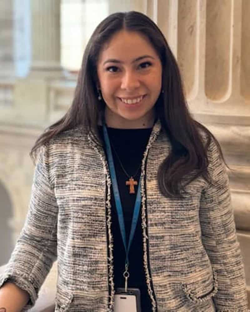 Angeles Ponpa-Avada-800×1000 Portrait of a smiling woman in a tweed blazer with lanyard and cross necklace.