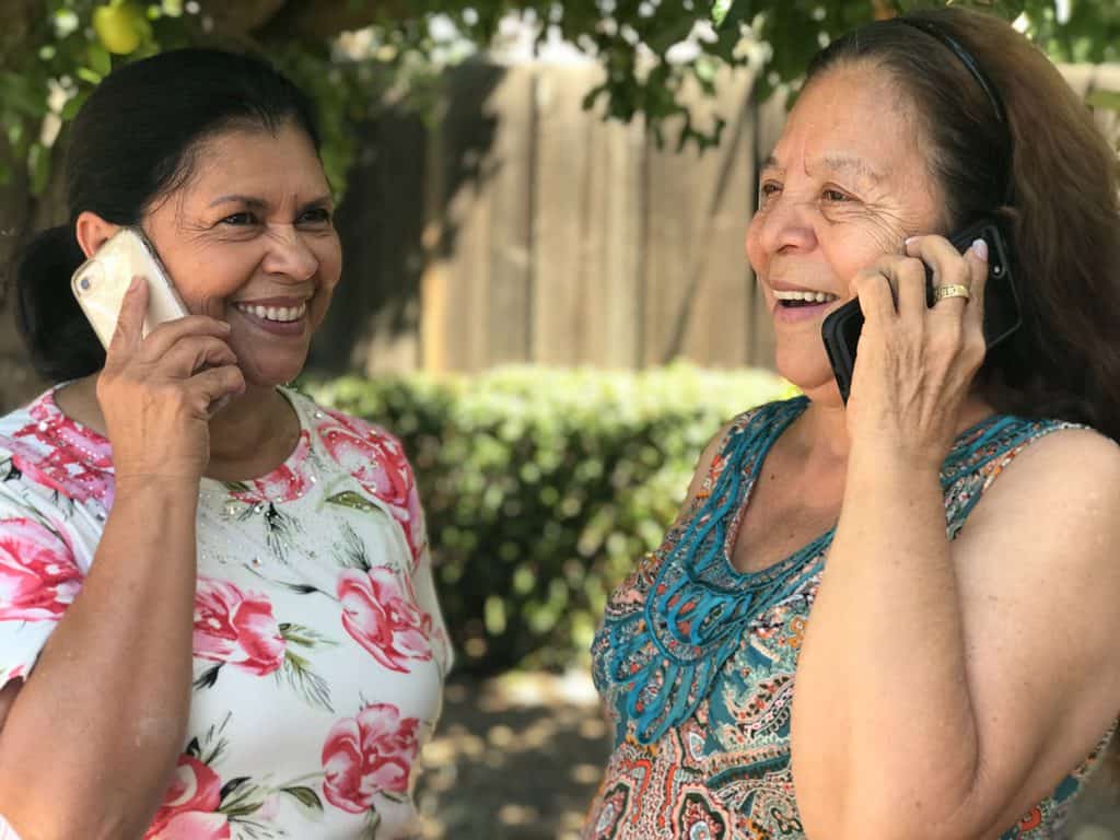 Two women talking on phones outdoors, smiling and enjoying each other's company.