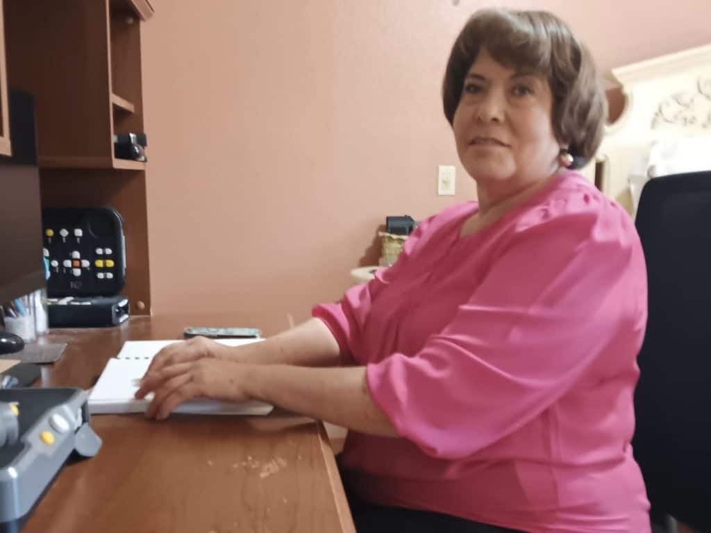 Woman with short dark hair in pink blouse reading Braille at desk.